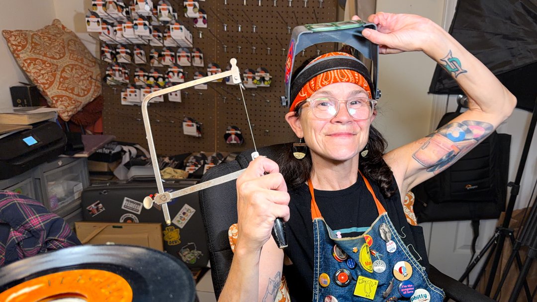 Leah Coonan at her jeweler's bench holding a jeweler's saw and visor, surrounded by vinyl records and handcrafted work